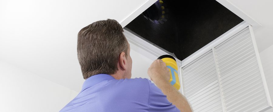 Male With A Yellow Flashlight Examining Hvac Duct System In A School